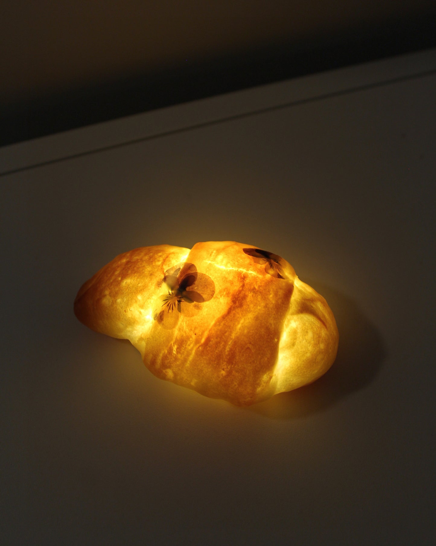 A mini bread roll lamp with daisies adorning it. Crafted from real, surplus bread that would otherwise be discarded-- baked, hollowed, and reimagined into a delightful lamp in Japan. Coated in an anti-bacterial and anti-fungal, eco-friendly resin.