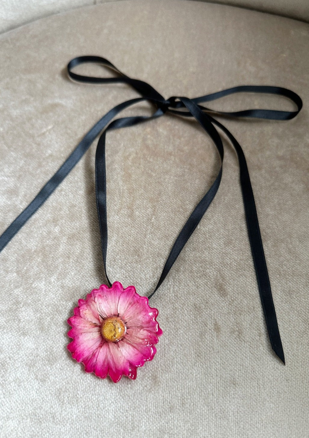 Pink daisy preserved in resin and suspended from a black satin ribbon.