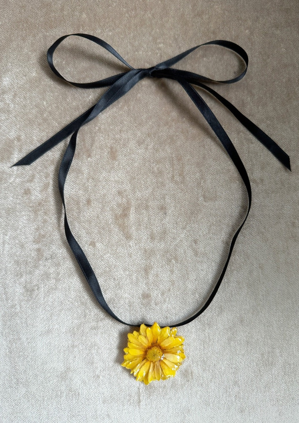 Yellow daisy preserved in resin and suspended from a black satin ribbon.
