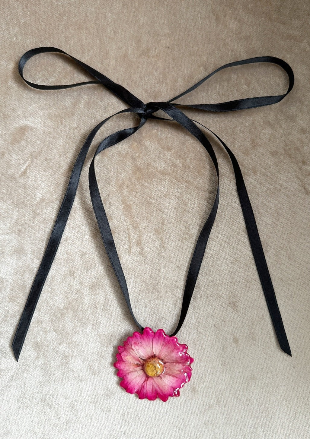 Pink daisy preserved in resin and suspended from a black satin ribbon.