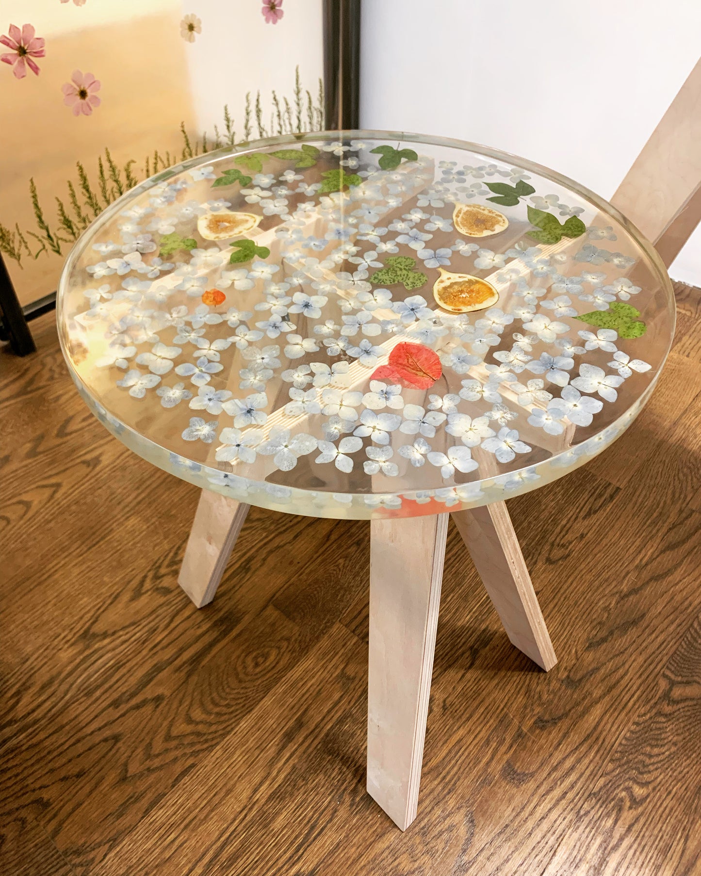 A clear round resin table top with blue hydrangeas, leaves, and figs scattered throughout. The table is held up by four light wooden legs